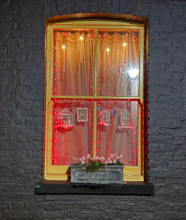 Lit window in dark wall, with hanging decorations of gingerbread houses, framed by red lights