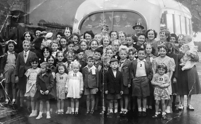 Black and white photo, 1950s, of coach with group of mostly children standing at the back of it, arranged for a photo. Probably going on an outing